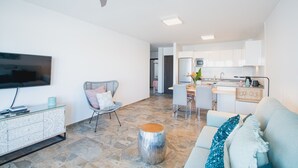 A modern living room with a flat-screen TV, a glass coffee table, a woven chair, and a sofa with decorative pillows. A dining area with a table and chairs is visible in the background. - Planta Baja - Villa Arosa (Playa Blanca)