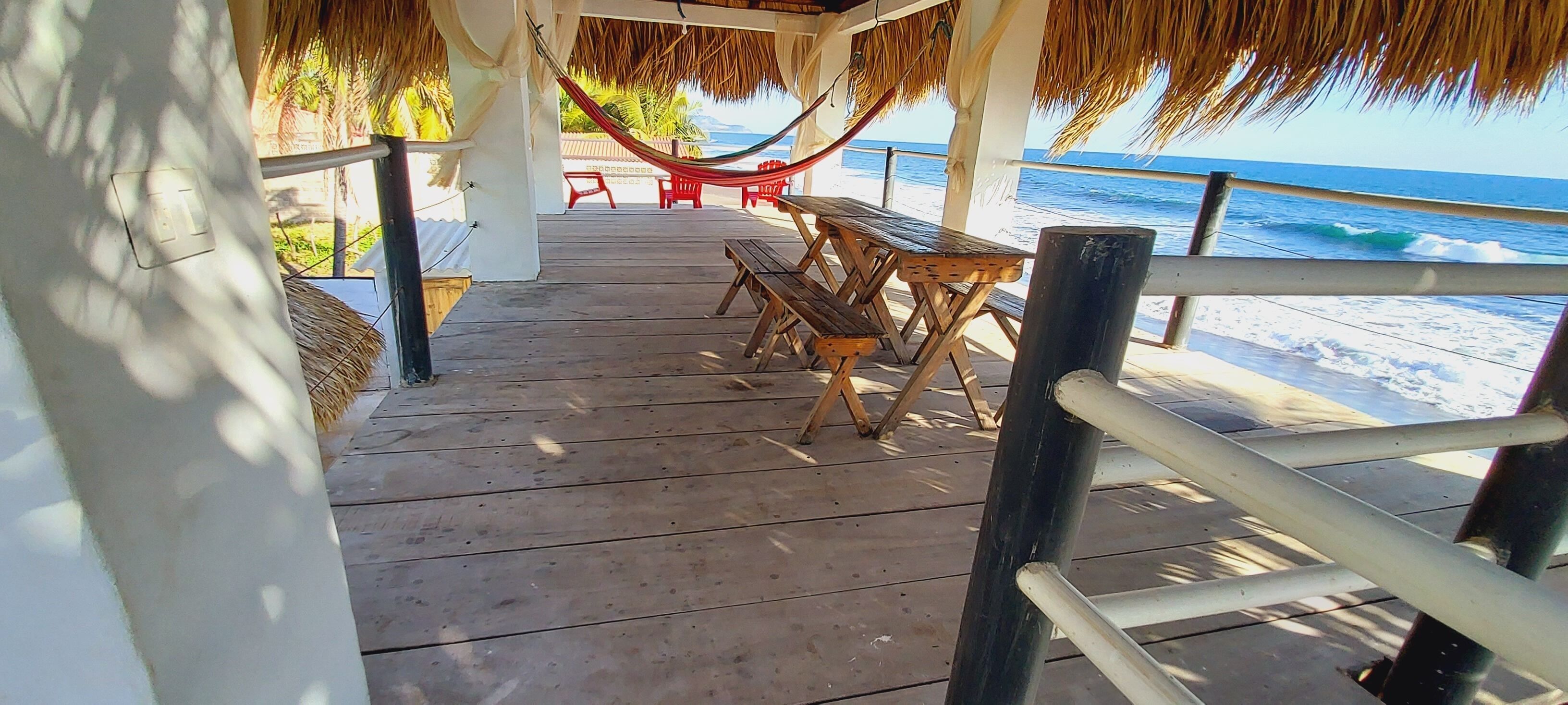 Une terrasse en bois avec une table et des chaises, un hamac et une rambarde d’où l’on a vue sur la plage.