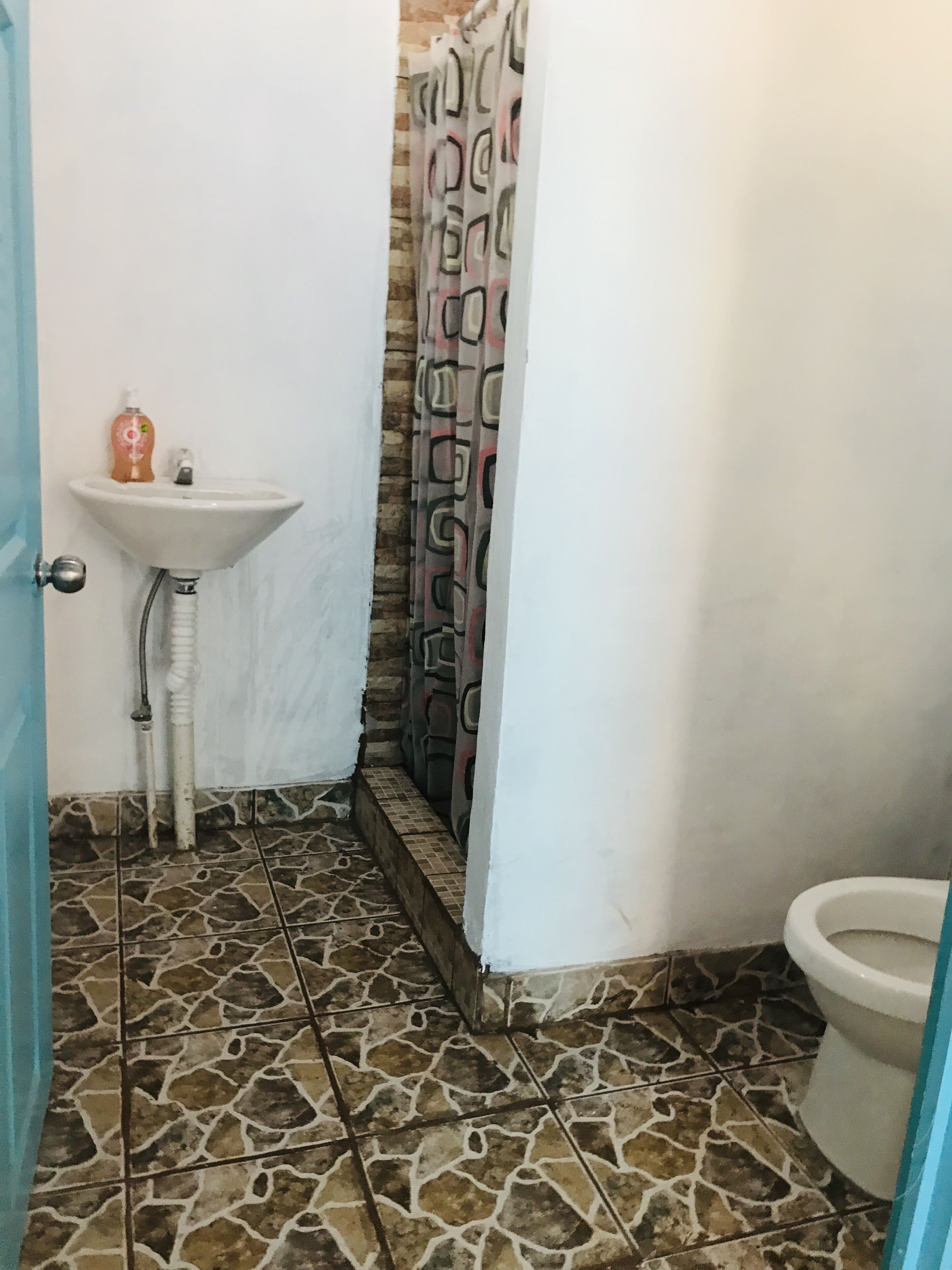 Um banheiro com piso de ladrilho de pedra, pia branca com dispensador de sabonete e cortina de chuveiro com padrão geométrico.
