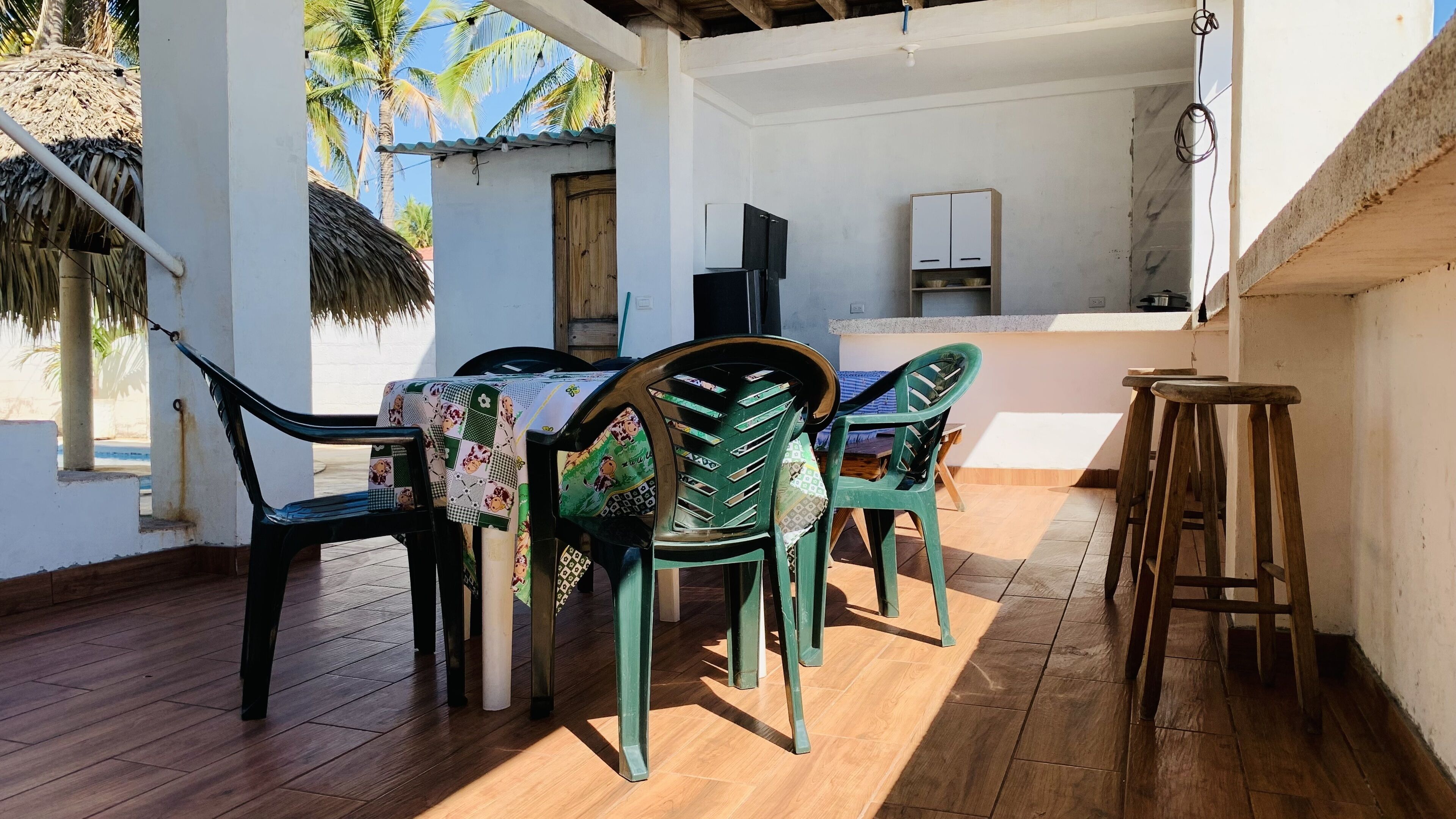 Une terrasse dotée d’un plancher en bois, de chaises en plastique vertes, d’une table recouverte d’une nappe à motifs, d’un tabouret en bois et d’un coin cuisine avec des placards et un réfrigérateur.