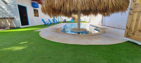 Una piscina circular con una fuente central, rodeada por una solana de hormigón y una zona ajardinada.