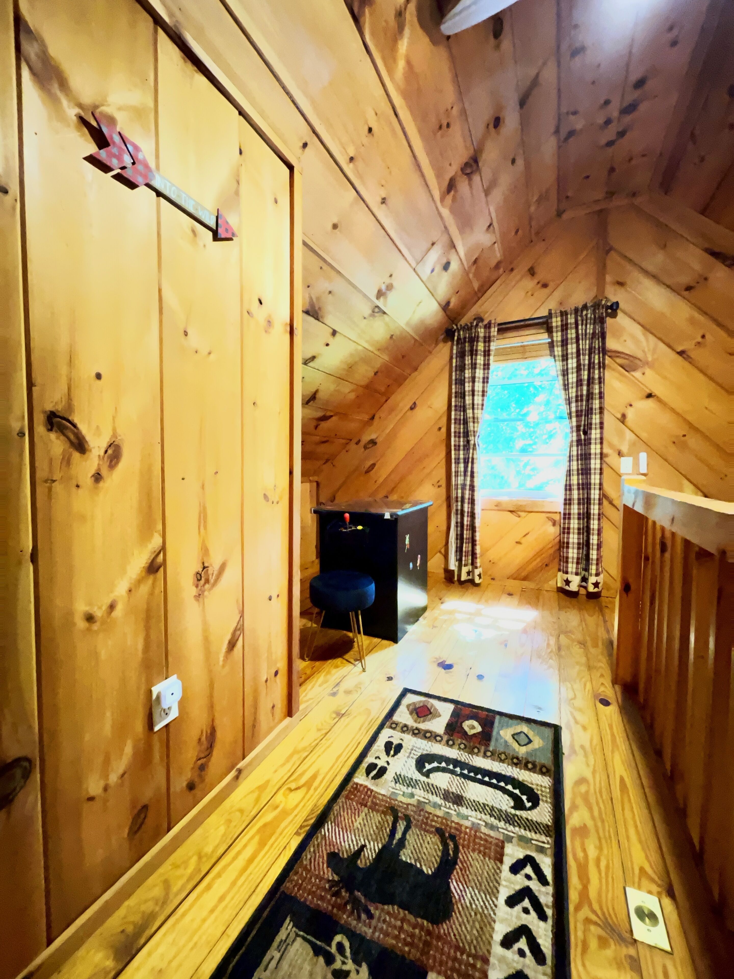A wooden interior with a sloped ceiling, a small seating area with a blue chair, a patterned rug, and a window with plaid curtains.