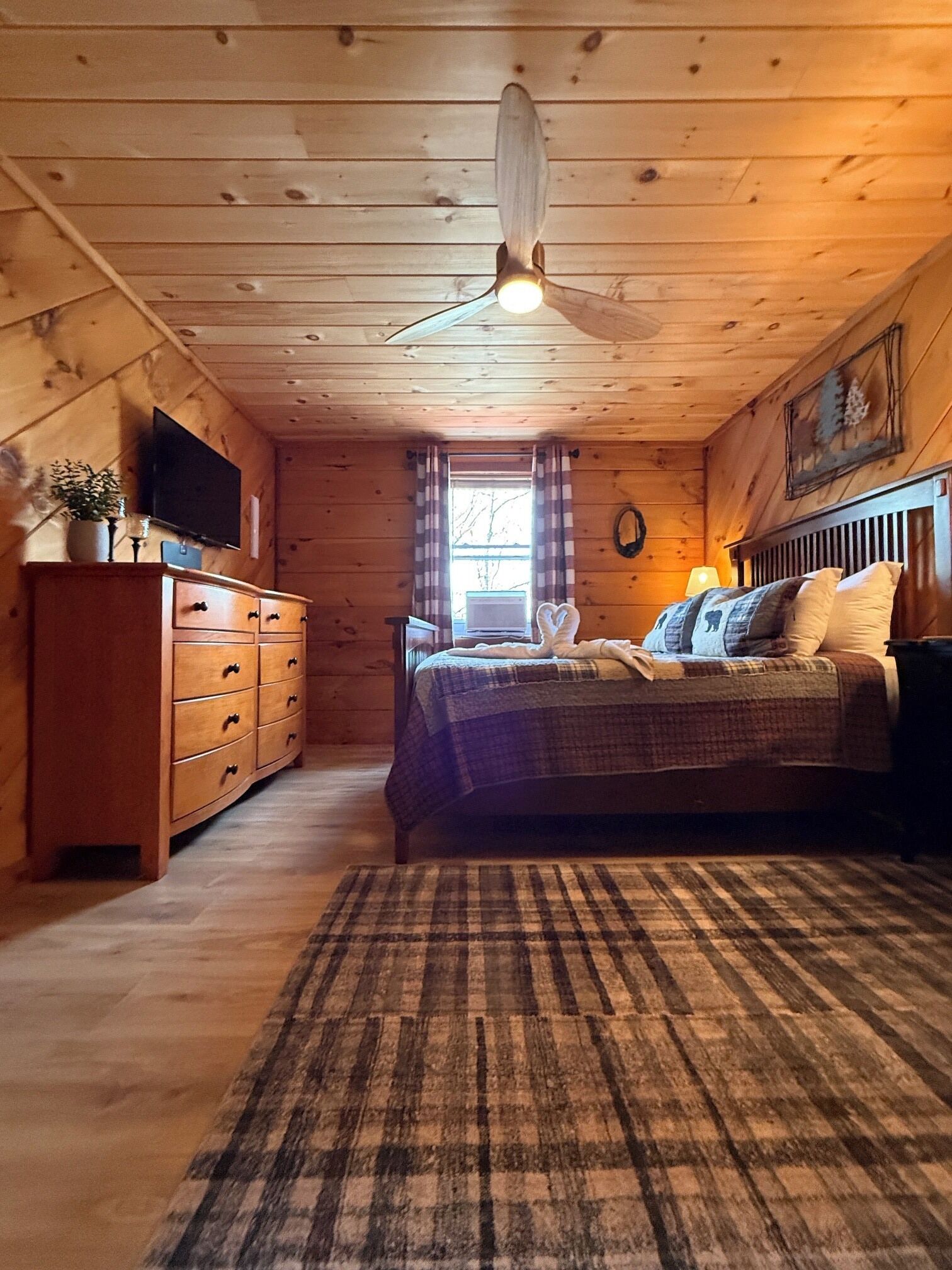 A cozy cabin bedroom with a wooden ceiling, a bed with plaid bedding, a dresser, a television, and a window with curtains.