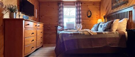A cozy cabin bedroom with a wooden ceiling, a bed with plaid bedding, a dresser, a television, and a window with curtains.