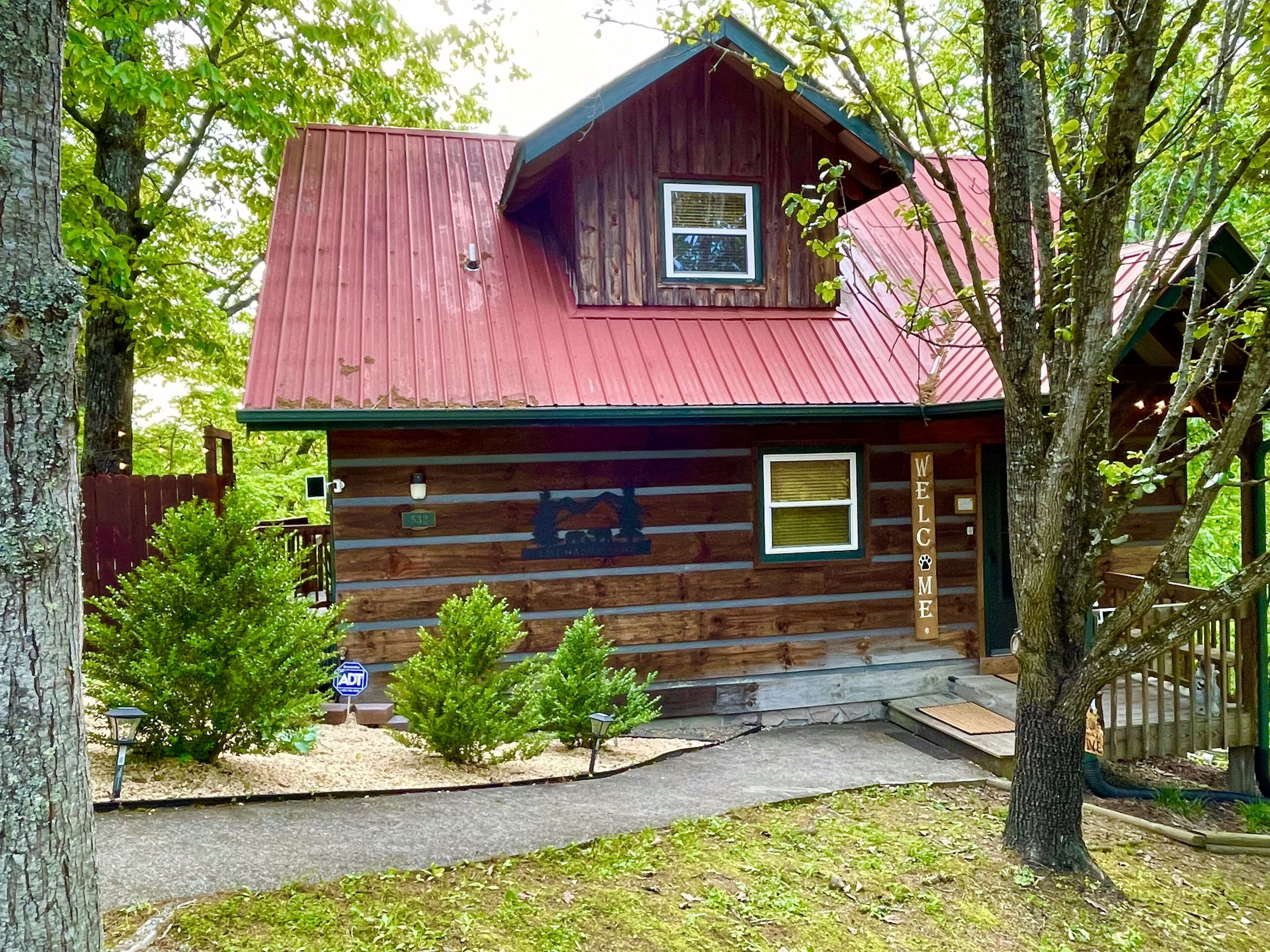A rustic log cabin with a red roof, a small porch, and a welcome sign.