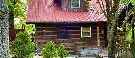 A rustic log cabin with a red roof, a small porch, and a welcome sign.