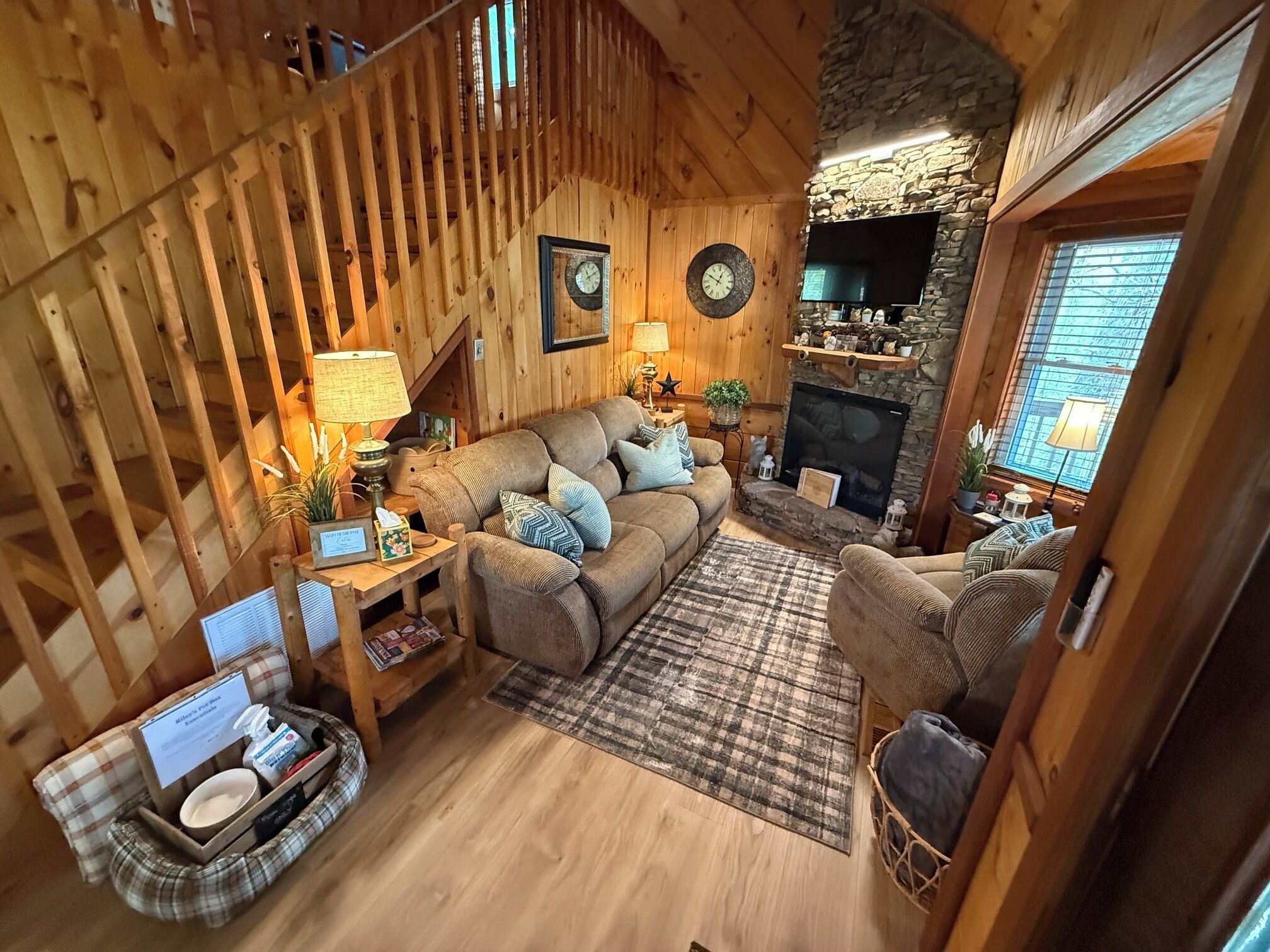 A cozy cabin interior with a wooden staircase, a fireplace, a sofa, and a television.