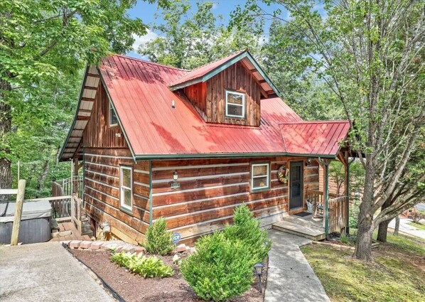 Une cabane rustique en rondins de bois, avec un toit en métal rouge, une véranda et un petit jardin.