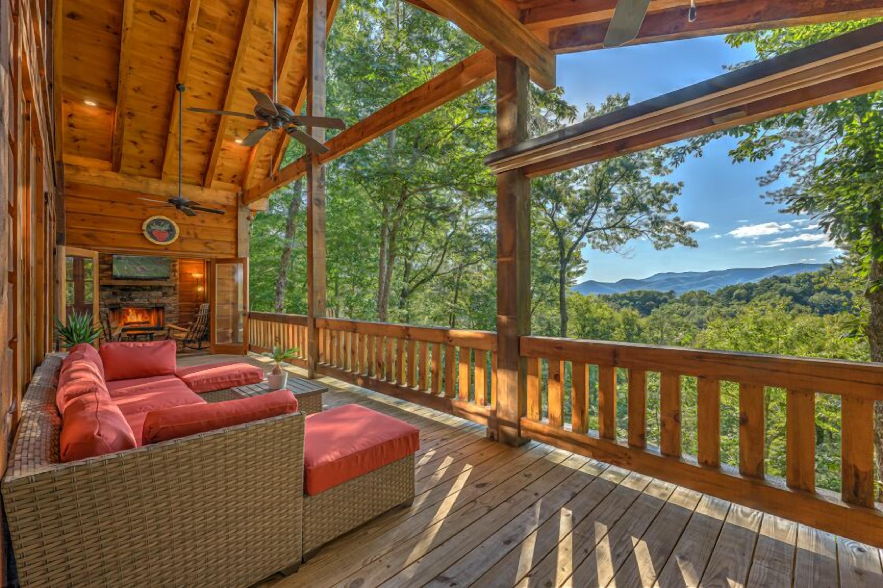 A wooden deck with a wicker sofa and red cushions, overlooking a forested landscape.