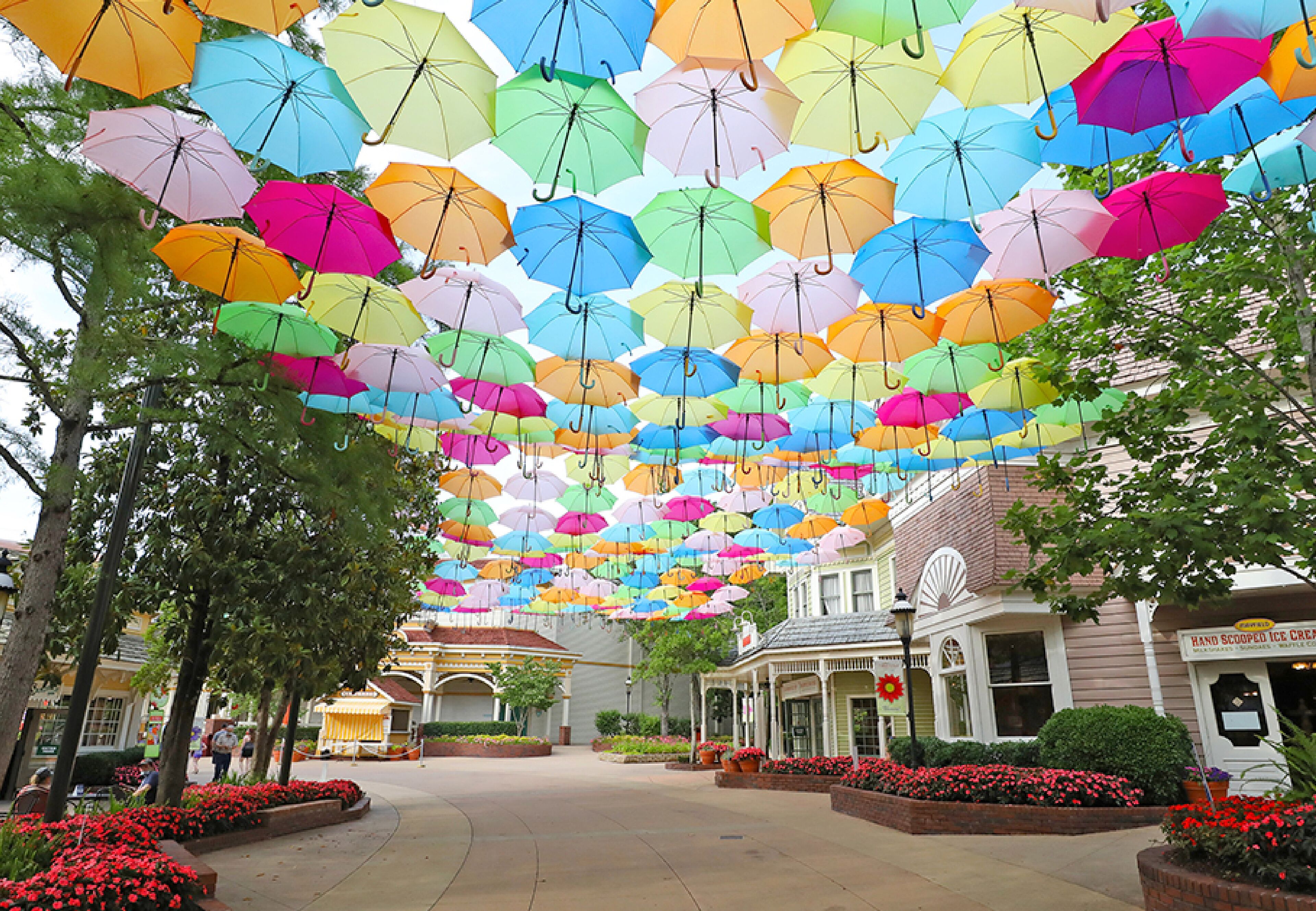 Una calle con paraguas de colores colgando, edificios y macizos de flores.