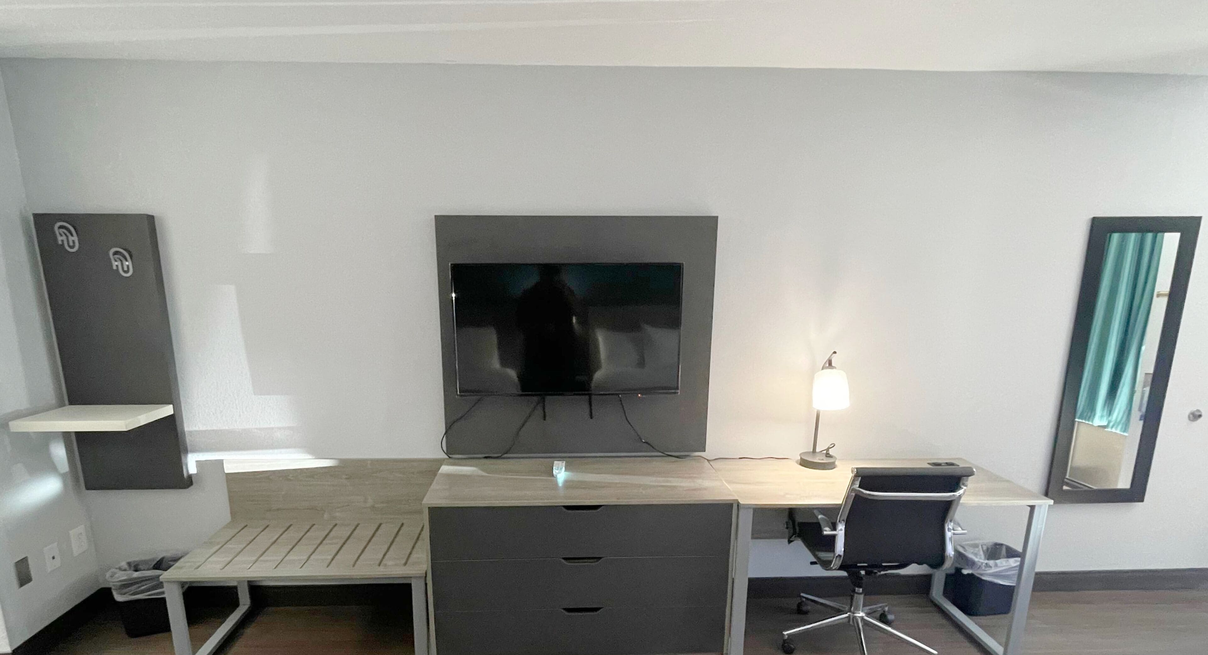 A modern hotel room with a flat-screen TV mounted on the wall, a desk with a chair, a small lamp, and a mirror.