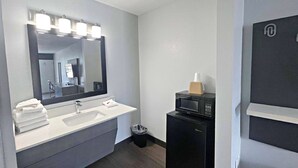 A modern bathroom with a white sink, a large mirror with lights, a trash bin, and a black microwave.
