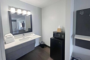 A modern bathroom with a white sink, a large mirror with lights, a trash bin, and a black microwave.