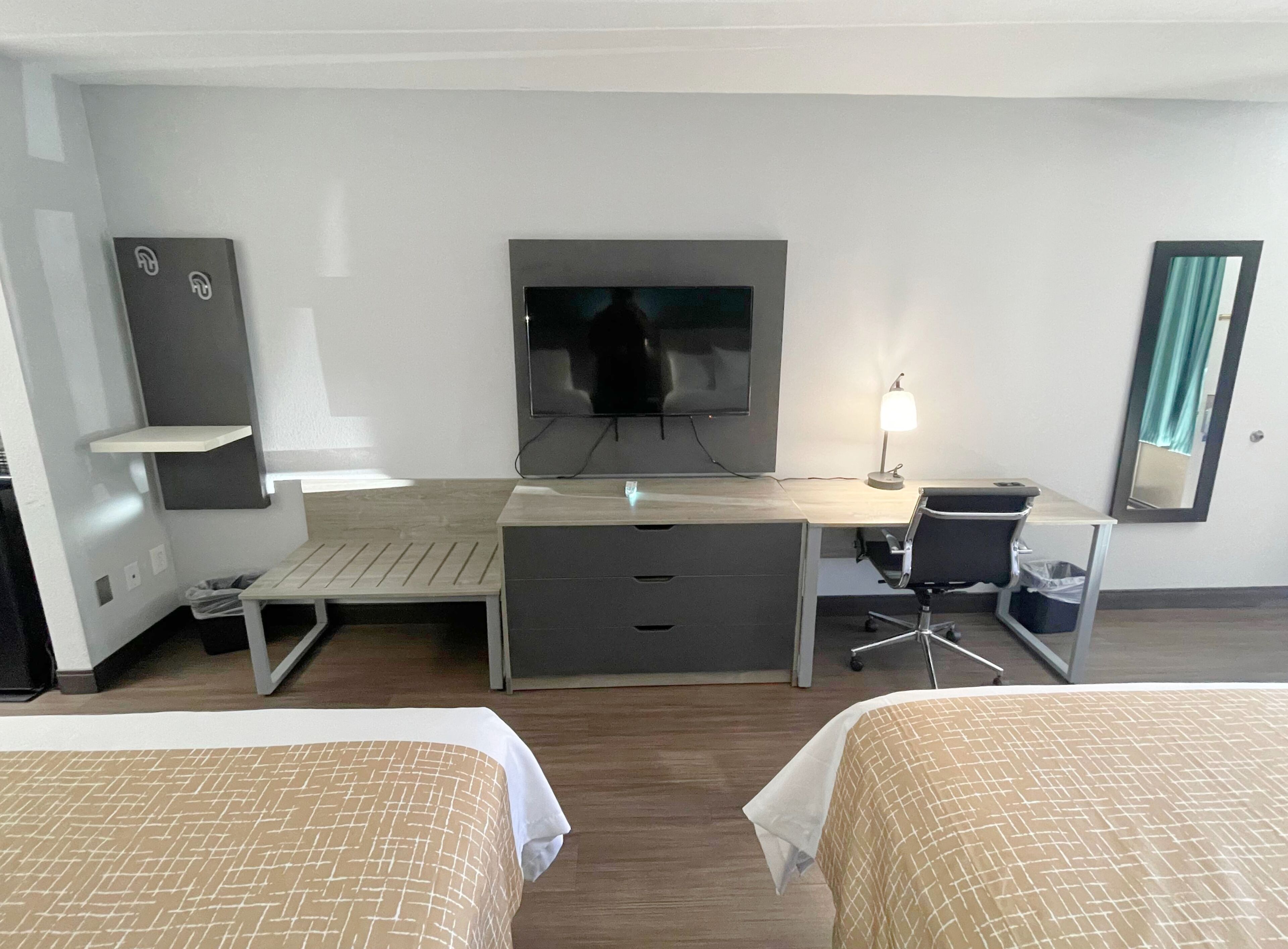 Habitación de hotel con dos camas, un escritorio con silla, televisor, lámpara, cesto de basura y espejo.