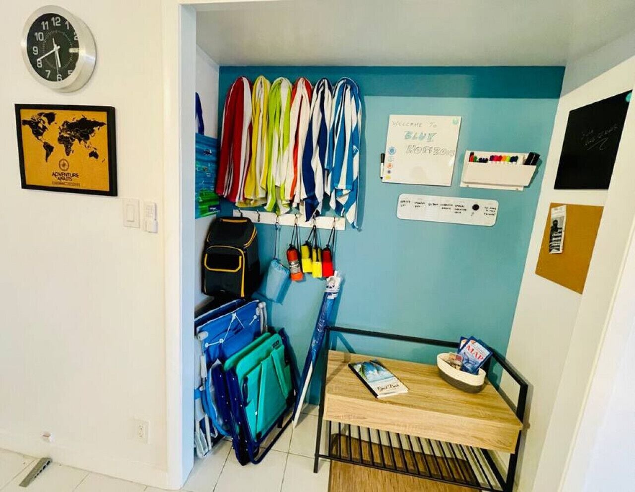 Een kleine kamer met een blauwe wand, een wereldkaart, een klok, en diverse spullen zoals een rugzak, kleding en een klein tafeltje met een schaal lekkernijen.