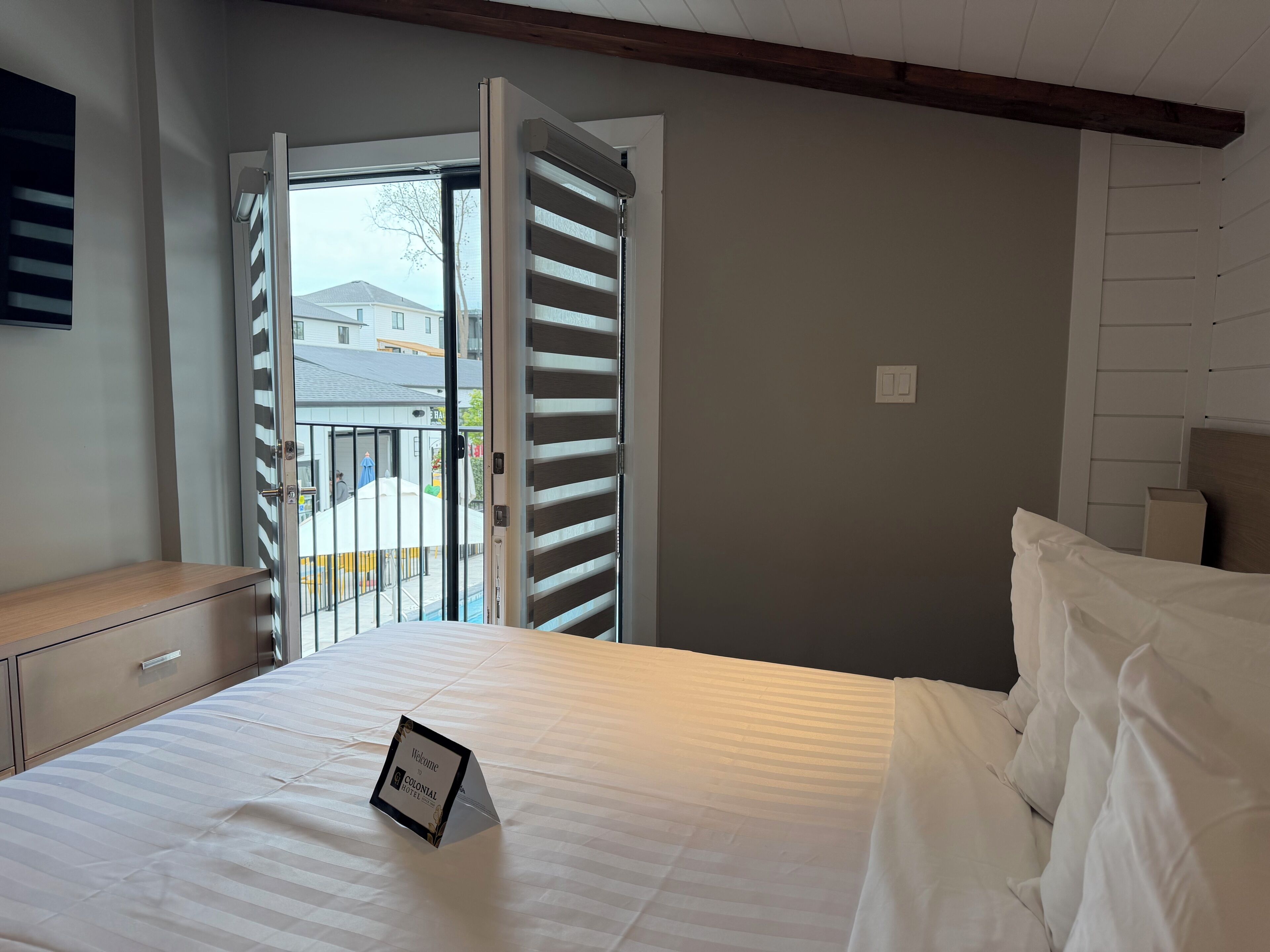 Habitación de hotel con cama, mesita de noche y ventana con persianas, con vistas al exterior.