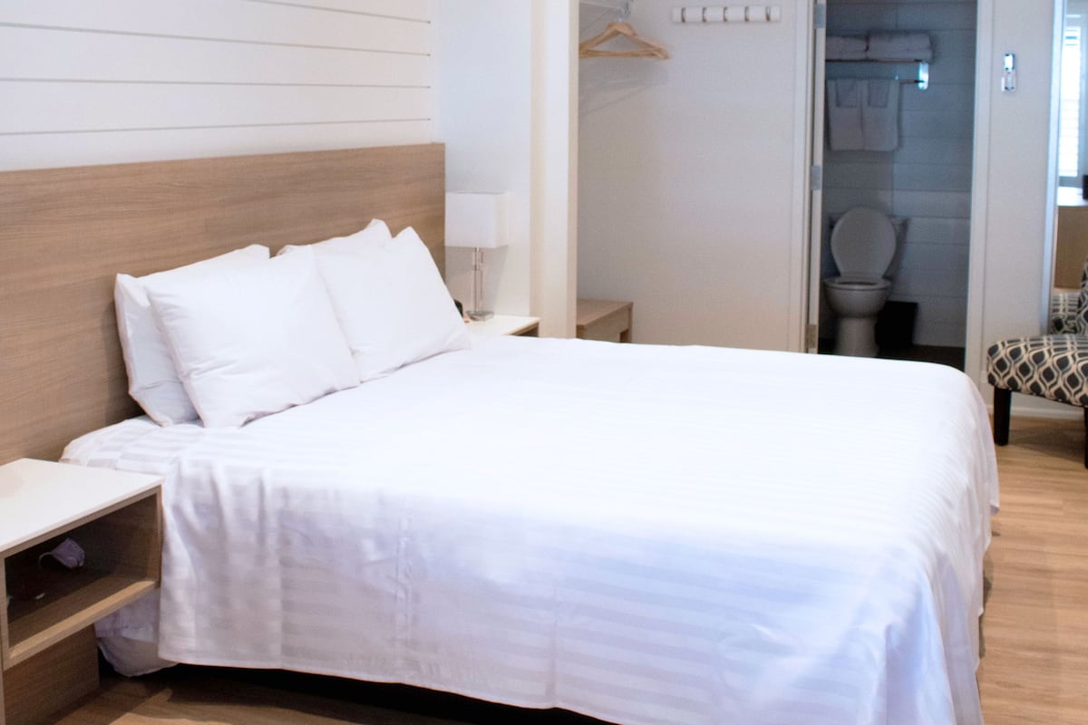 Un lit bien fait, avec une tête de lit en bois, une table de chevet et une chaise agrémentée d’un coussin à motifs.