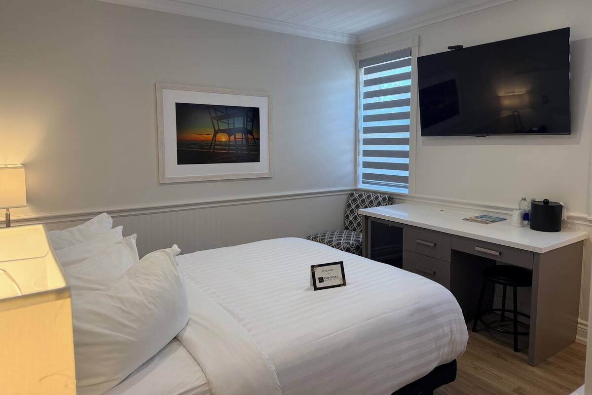 Une chambre d’hôtel avec un lit, un bureau, une télévision, un tableau encadré et une fenêtre avec des stores.