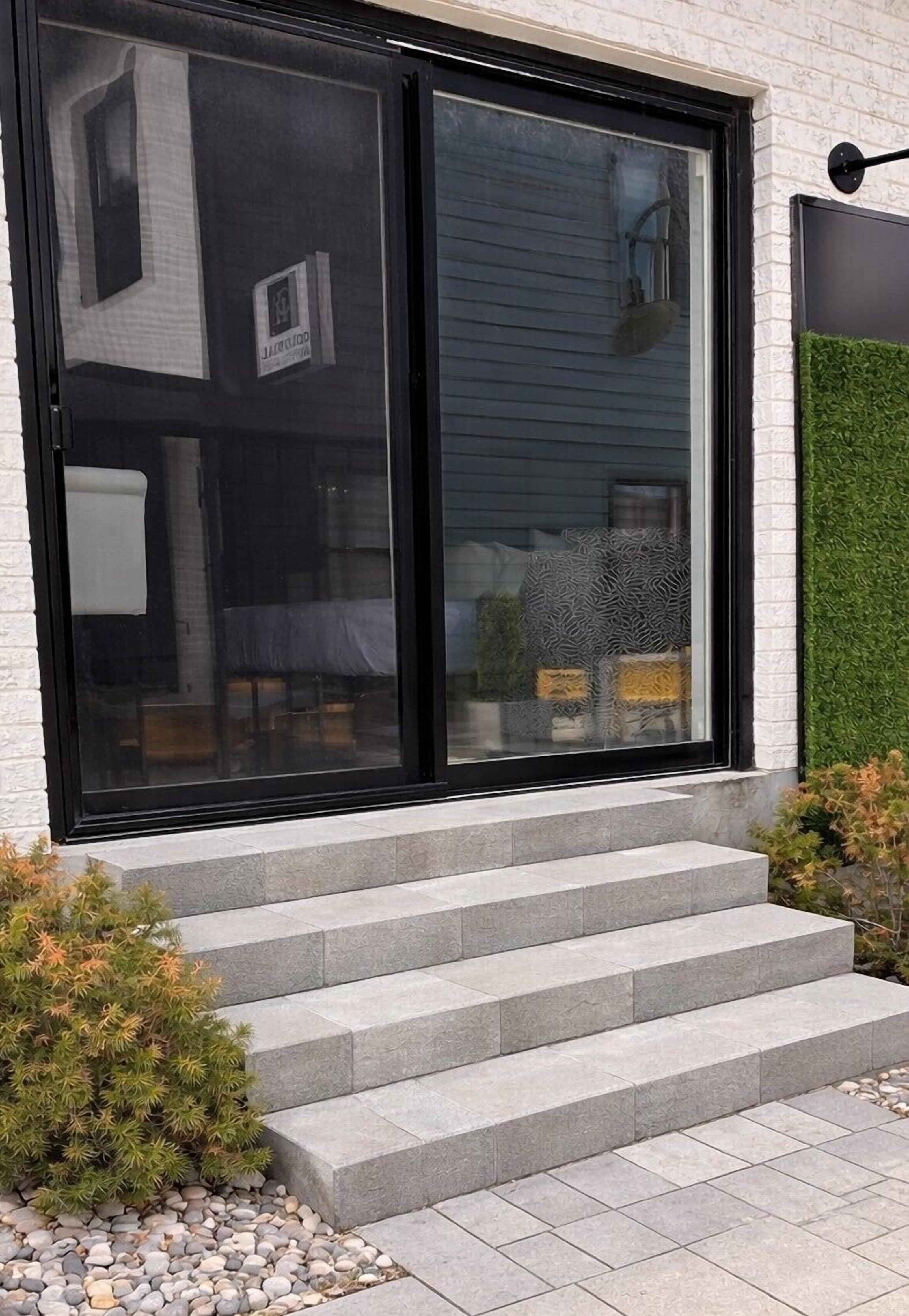 Un edificio moderno con grandes puertas correderas de vidrio, una escalinata de piedra y un pequeño jardín con arbustos.