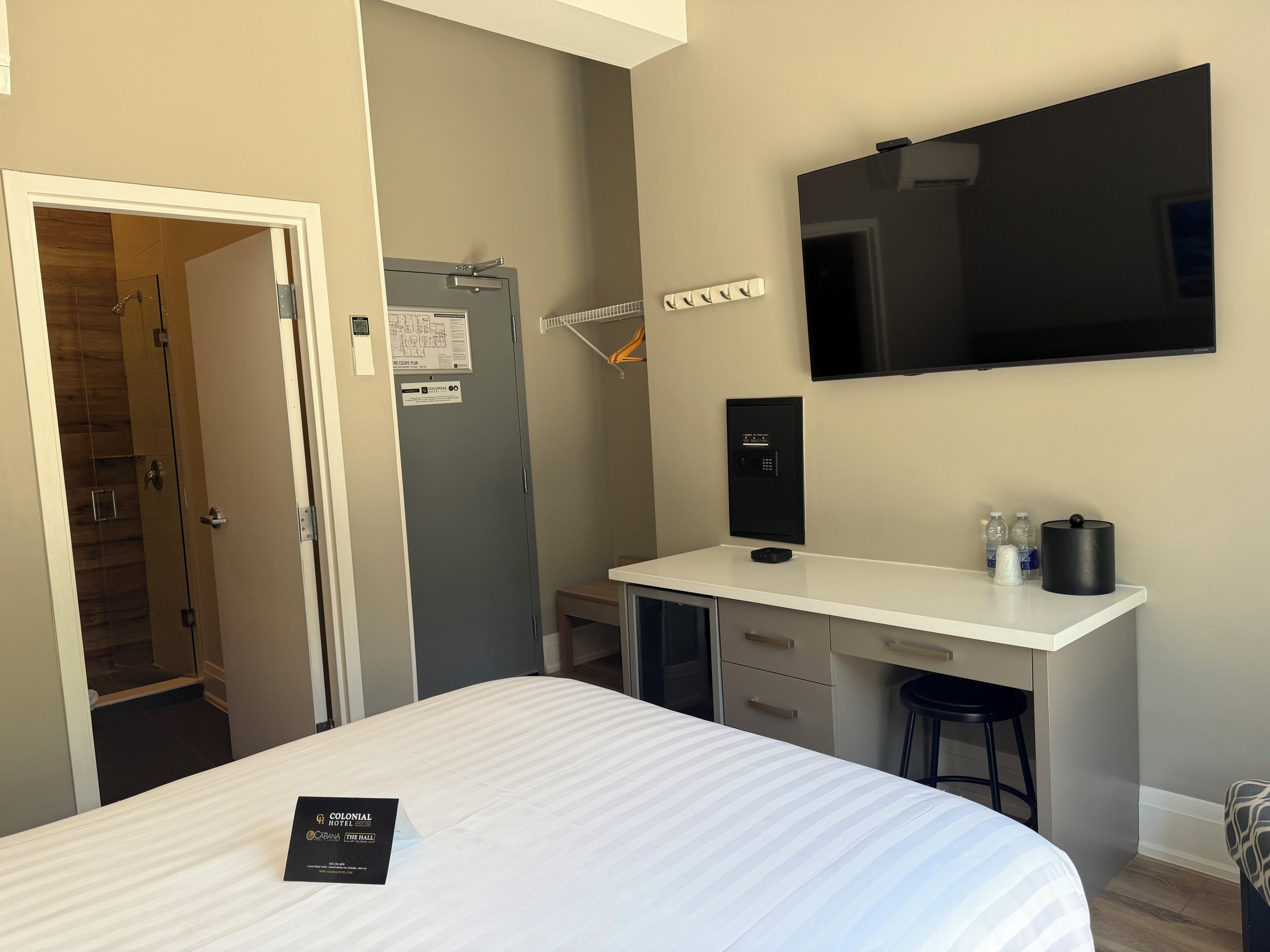A hotel room with a bed, a desk, a chair, a TV, a safe, and a door leading to a shower.