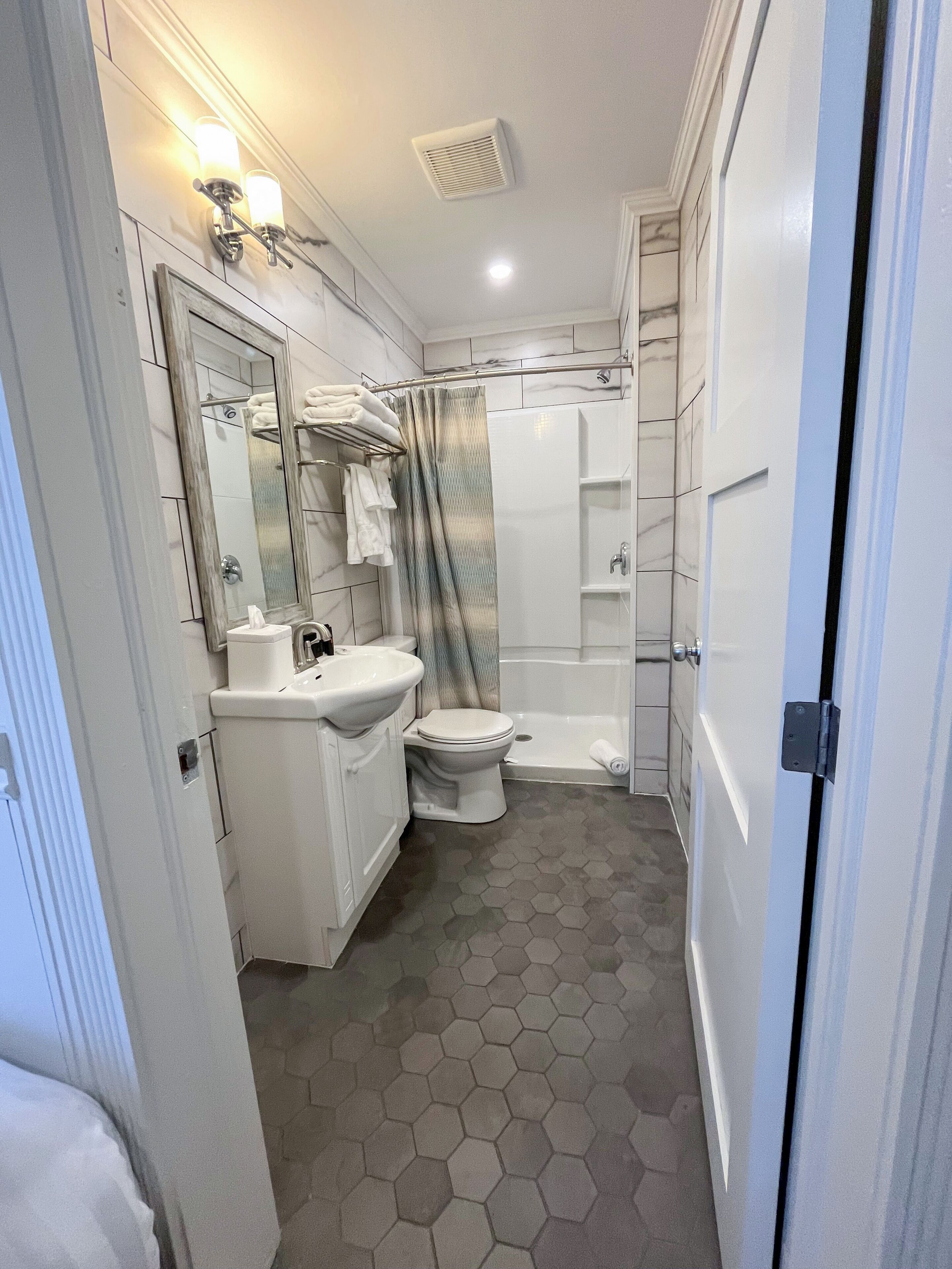 Une salle de bain avec un lavabo, des toilettes et une douche blancs, un miroir et un sol carrelé avec des carreaux hexagonaux.