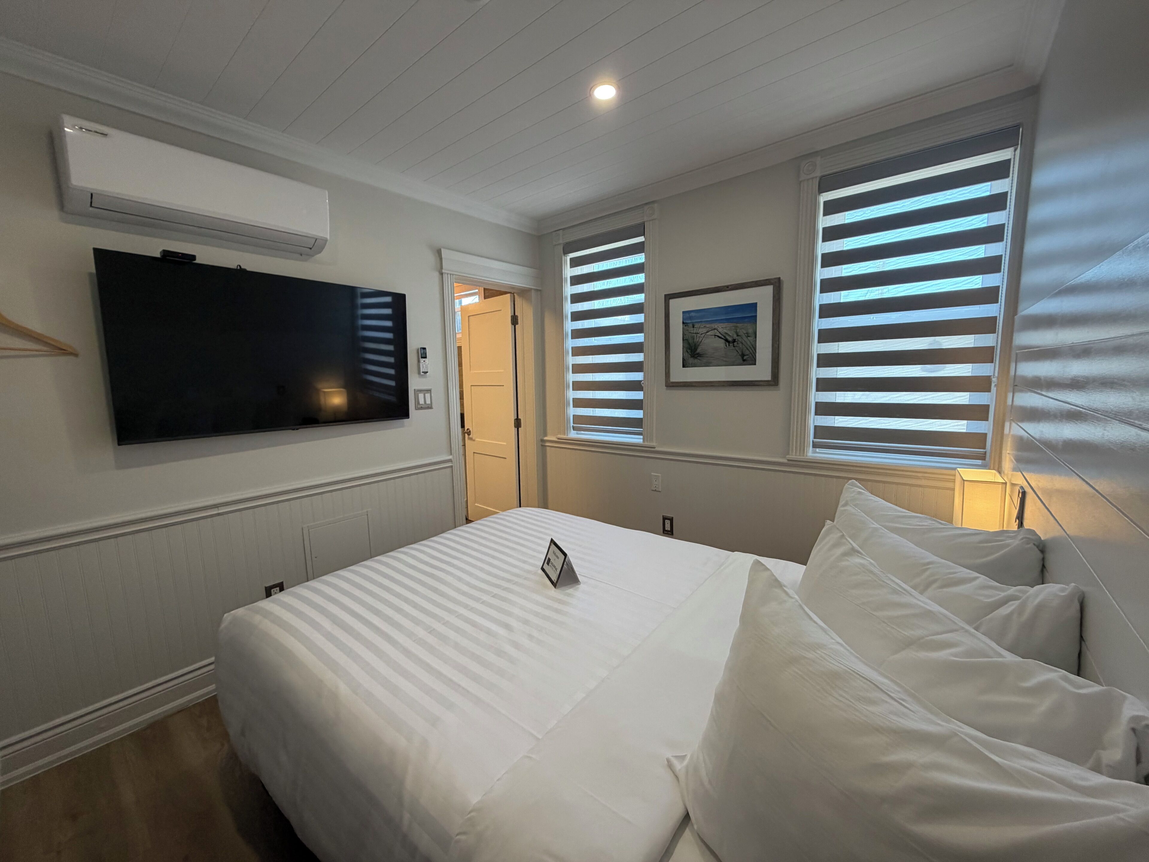 Habitación de hotel con cama, televisor de pantalla plana, un cuadro en la pared y ventana con persianas.