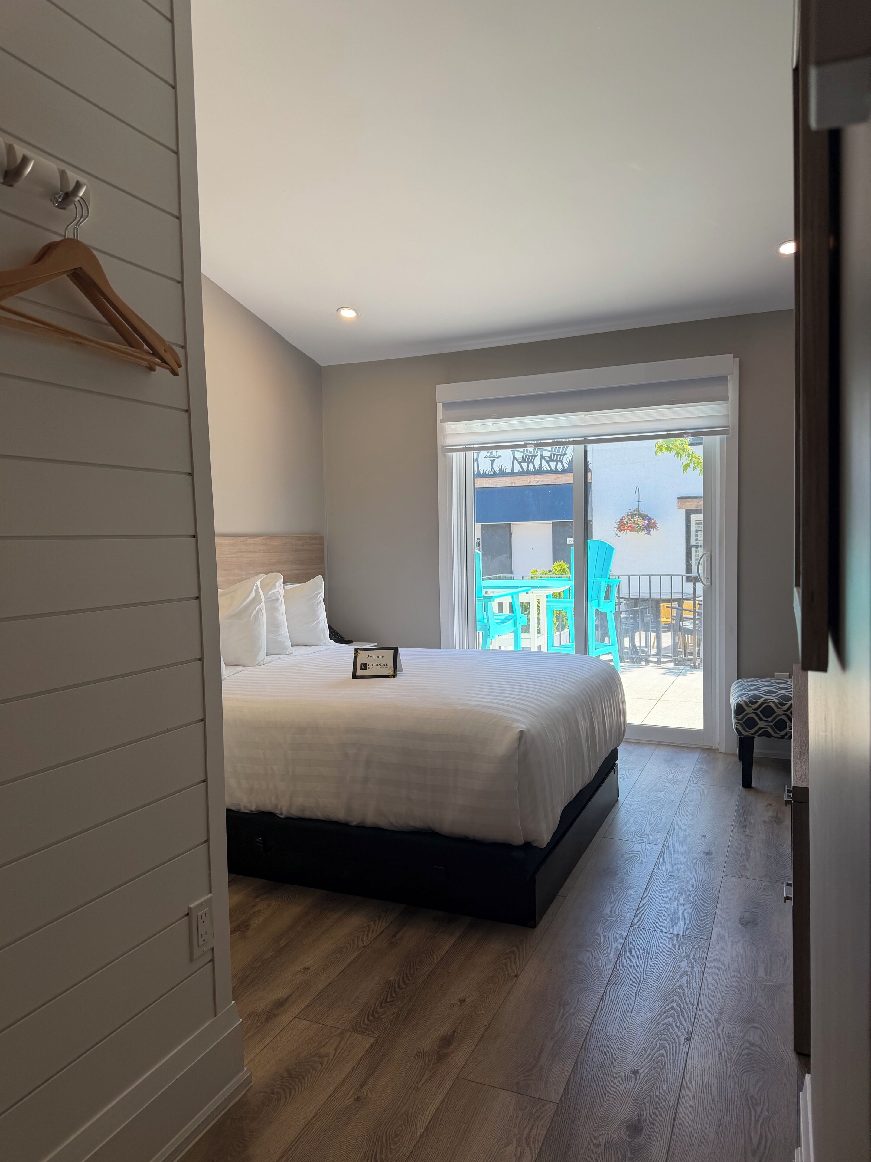 A modern hotel room with a large bed, a wooden floor, a sliding glass door leading to an outdoor area with chairs and a table.
