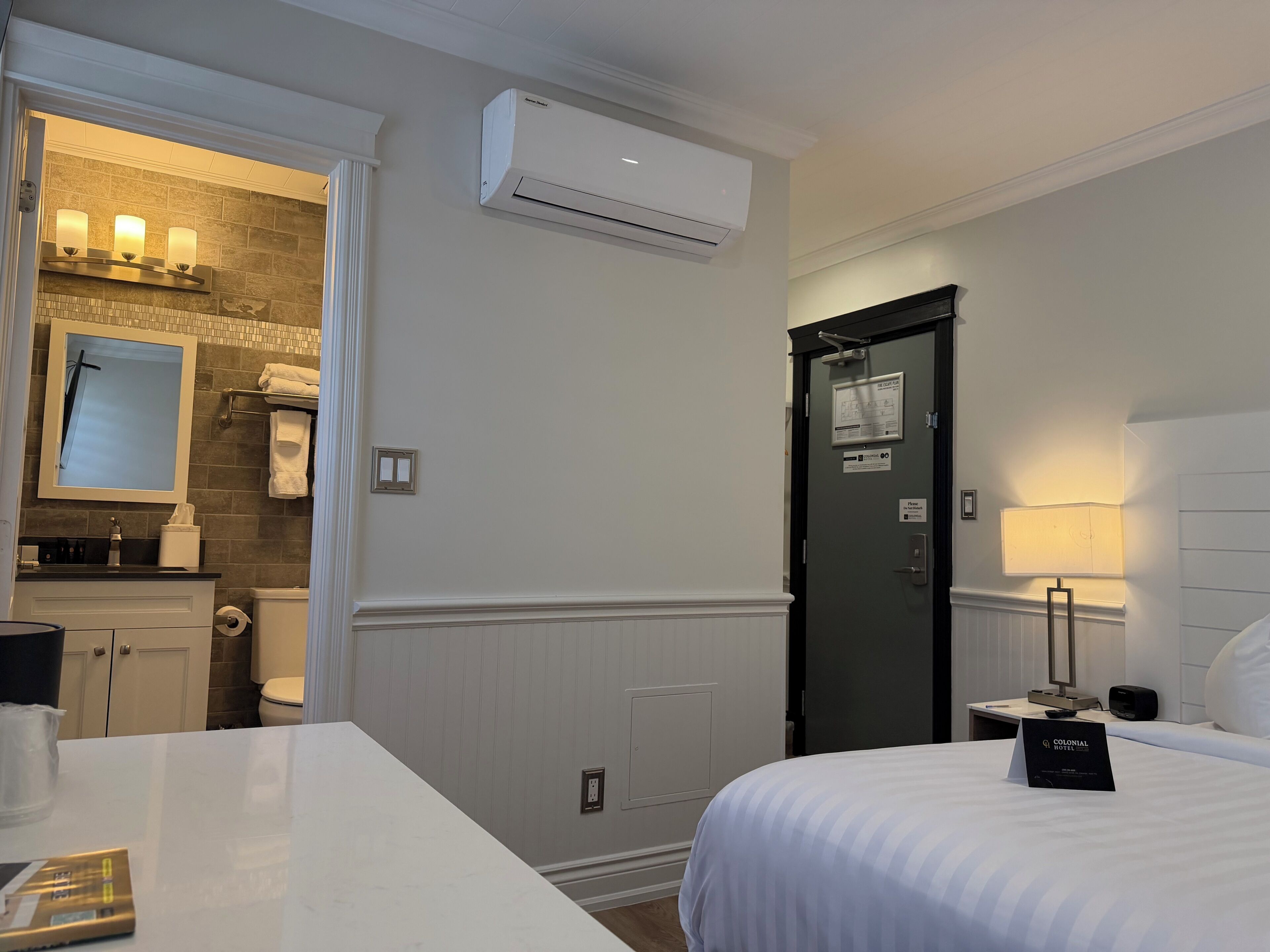 A hotel room with a bed, bedside table, lamp, air conditioning unit, and a bathroom with a mirror, sink, and towel rack.