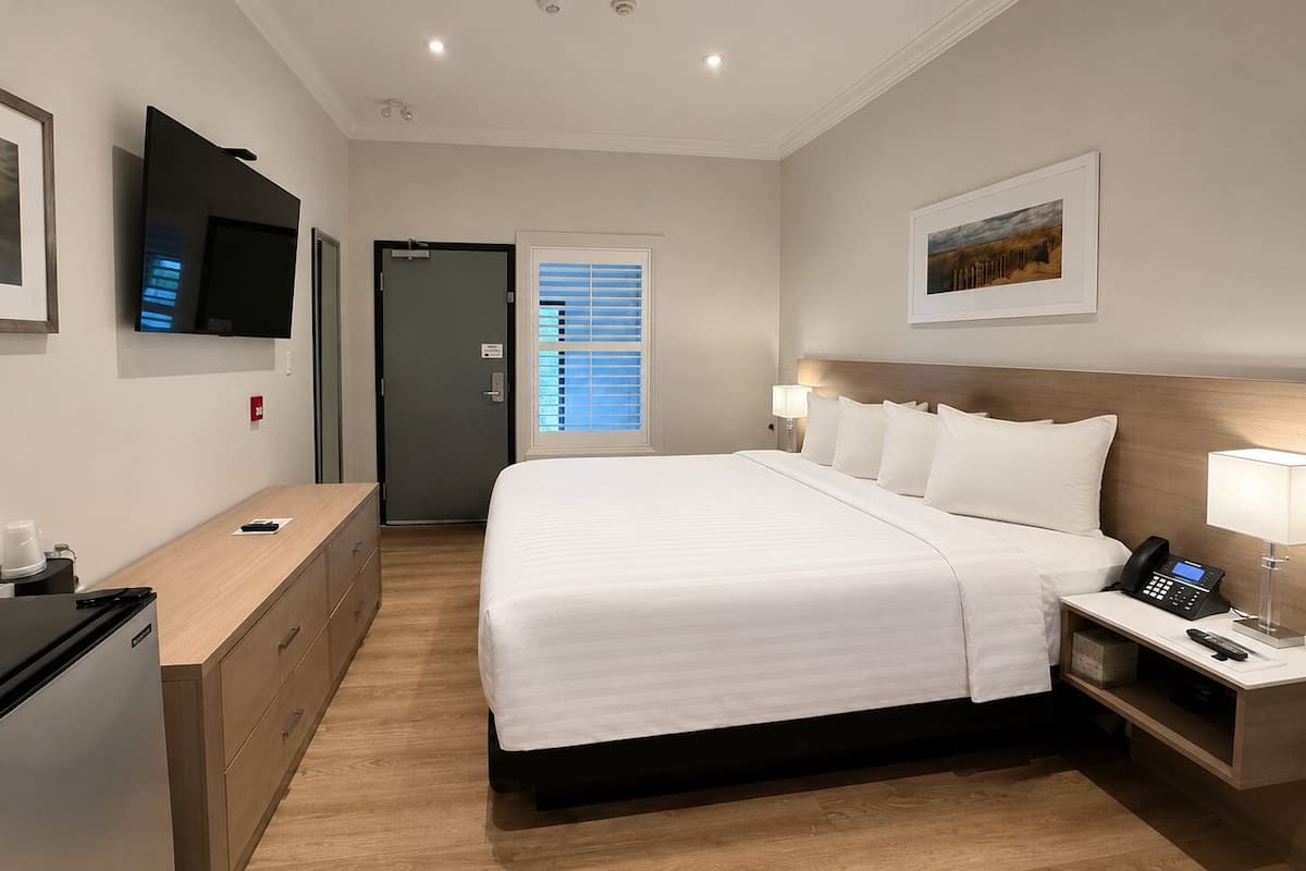 Une chambre d’hôtel comprenant un lit, une table de chevet avec un téléphone, un téléviseur fixé au mur, un bureau avec un ordinateur et une porte avec une fenêtre.