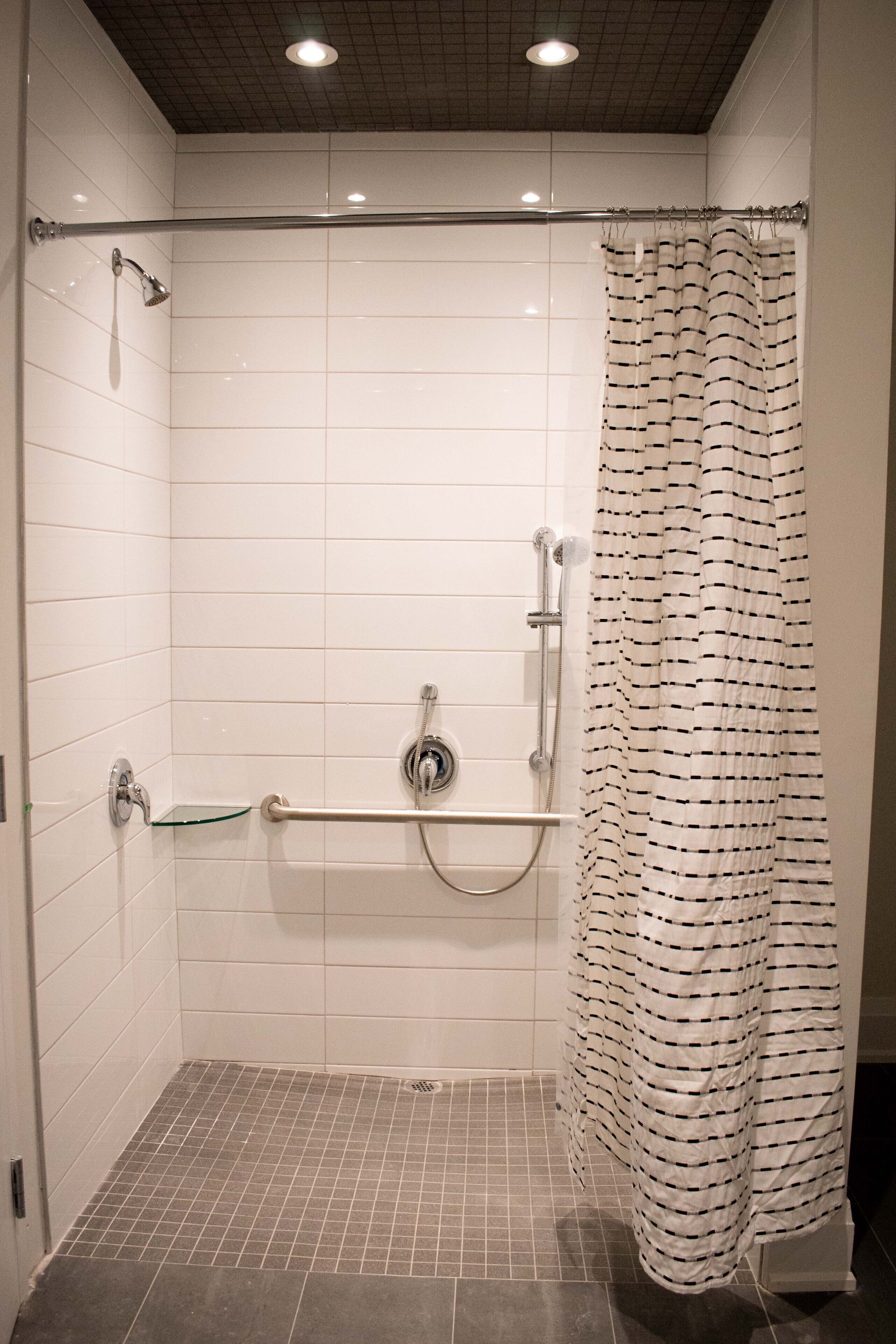 A shower area with white tiled walls, a glass shelf, a showerhead, a hand-held shower, a grab bar, and a striped shower curtain.