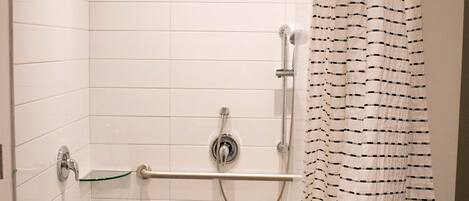 A shower area with white tiled walls, a glass shelf, a showerhead, a hand-held shower, a grab bar, and a striped shower curtain.