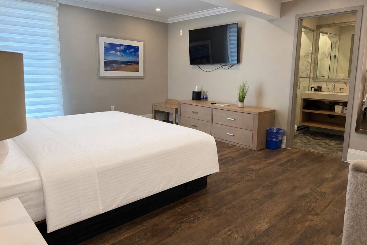 Une chambre à coucher avec un lit, une télévision, une commode, un miroir et une salle de bain visible à travers une porte ouverte.