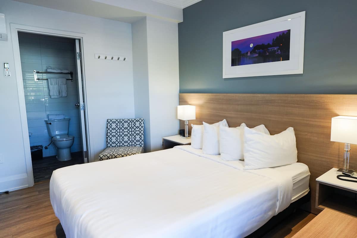 Une chambre d’hôtel avec un grand lit, deux tables de chevet avec des lampes, une petite table avec un téléphone, un tableau encadré au mur et une salle de bain visible à travers une porte ouverte.