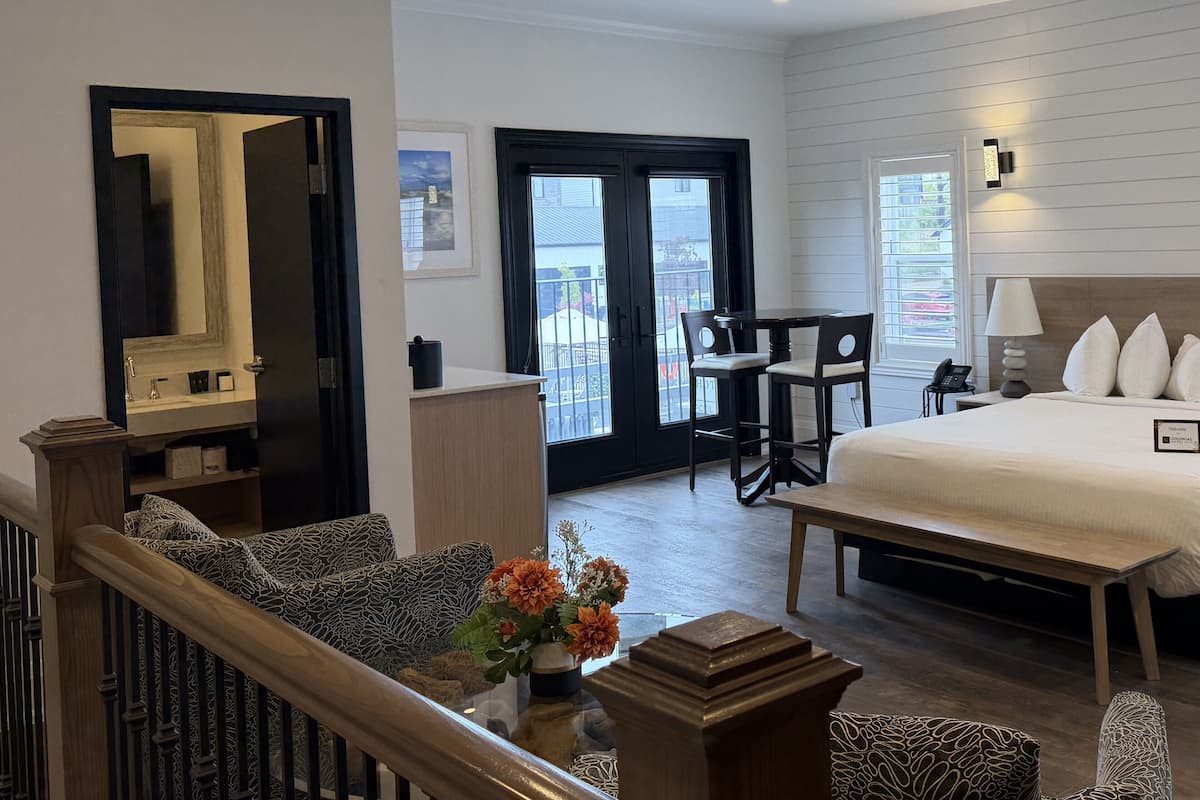 Une chambre d’hôtel comprenant un lit, un bureau, une chaise, une petite table, un miroir, une fenêtre et un escalier.