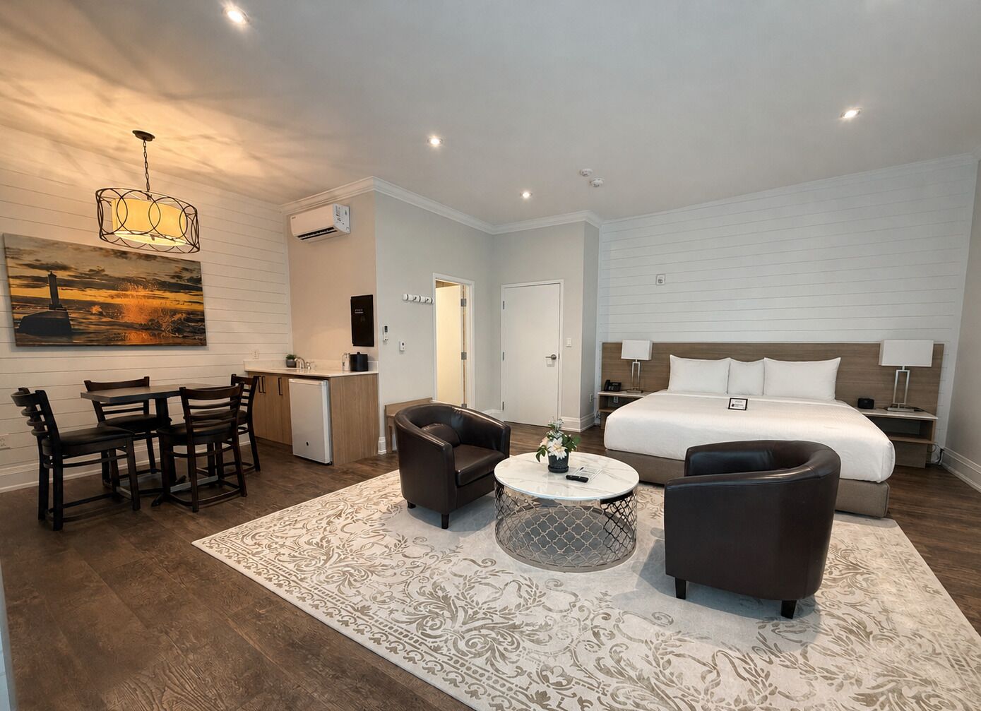 A modern hotel room with a bed, two armchairs, a small table, a dining area with chairs, a kitchen area, and a painting on the wall.