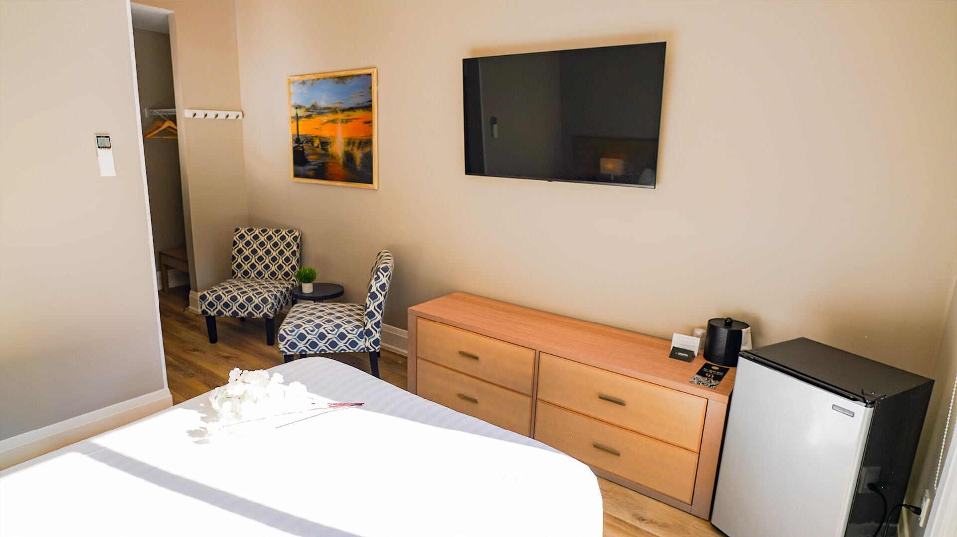 Une chambre d’hôtel comprenant un lit, deux fauteuils à motifs, une petite table avec une plante, une commode en bois, un téléviseur et un réfrigérateur.