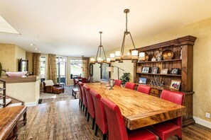 A spacious dining area with a long wooden table and red upholstered chairs, a wooden bookshelf with decorative items, and a view of a landscaped outdoor area through large windows.