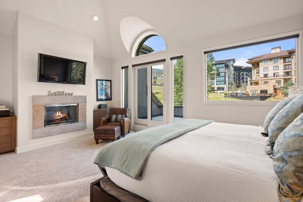 A bedroom with a fireplace, a large bed, a TV mounted on the wall, and a view of a building complex.