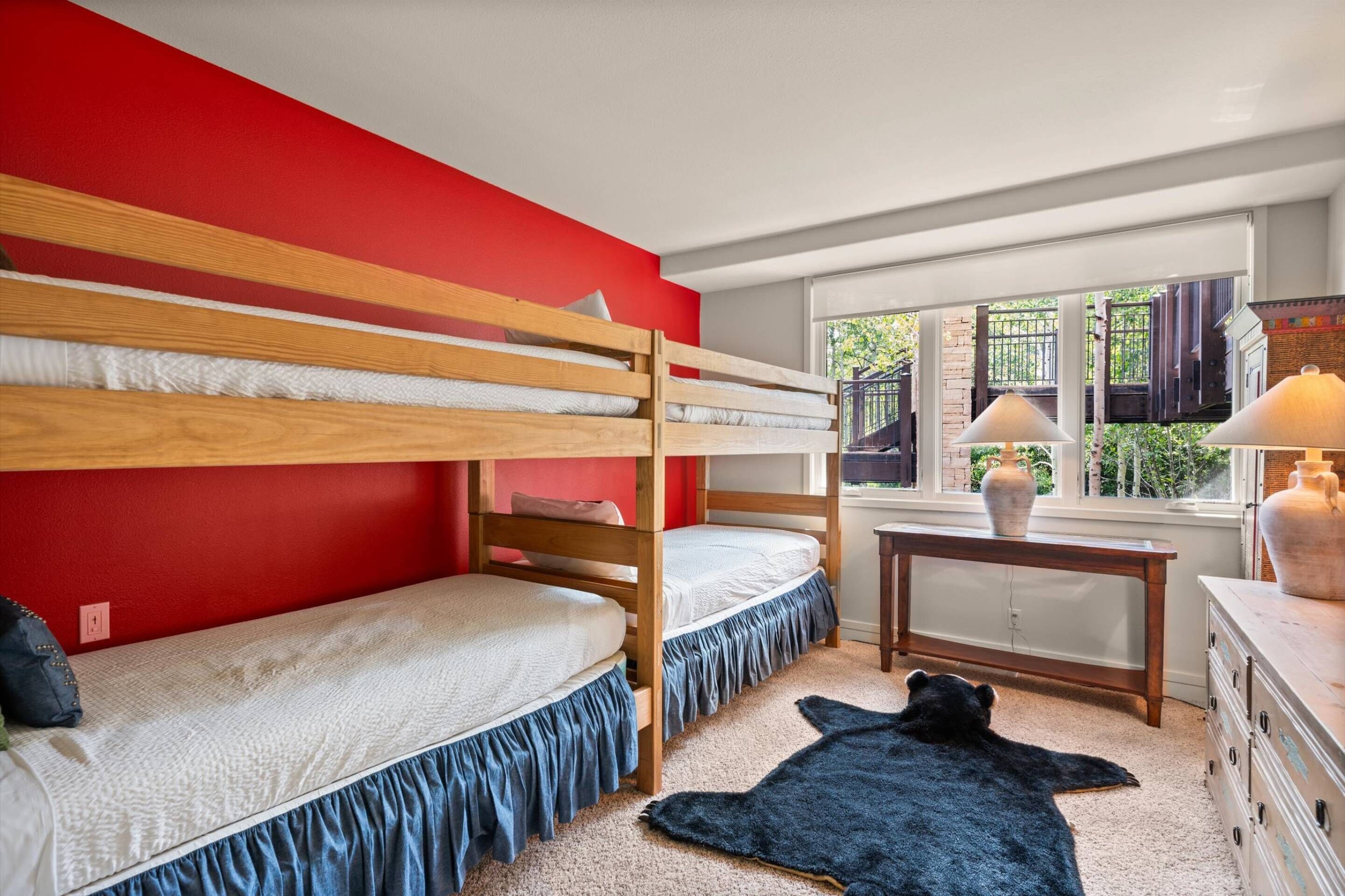 A room with two bunk beds, a window with a view of greenery, a small table with a lamp, and a dresser.