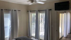 Une chambre avec un lit, un ventilateur de plafond, un téléviseur fixé au mur et une porte-fenêtre coulissante donnant sur un balcon.