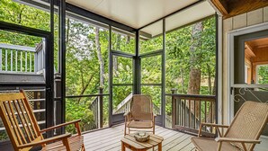 Um deck de madeira com cadeiras de vime e uma pequena mesa, com vista para uma floresta verde exuberante.