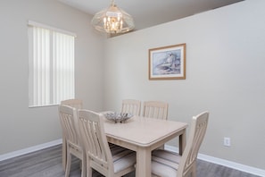 Une salle à manger avec une table et des chaises, un lustre, une fenêtre avec des stores et un tableau encadré au mur.
