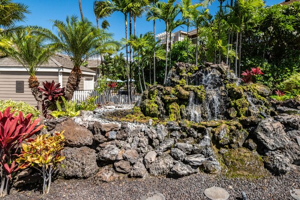 A tropical garden with a waterfall, palm trees, and a building in the background. - Serene Luxury Golf & Tennis Villa - outdoor pools, hot tub, bbq (Wailea-Makena)