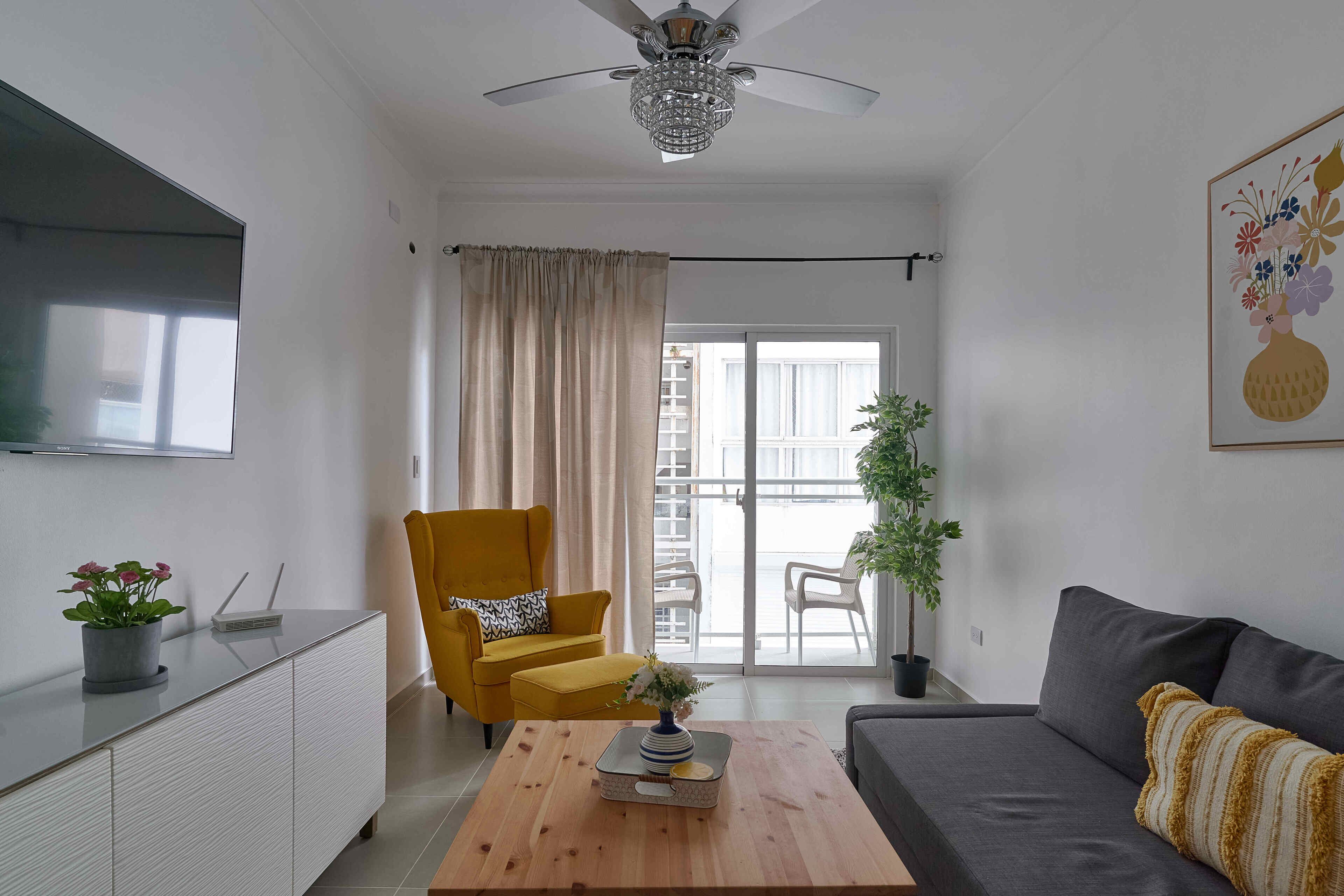 Un salon moderne comprenant un canapé, un fauteuil jaune, une table basse en bois, un ventilateur de plafond et une porte-fenêtre coulissante donnant sur un balcon extérieur.