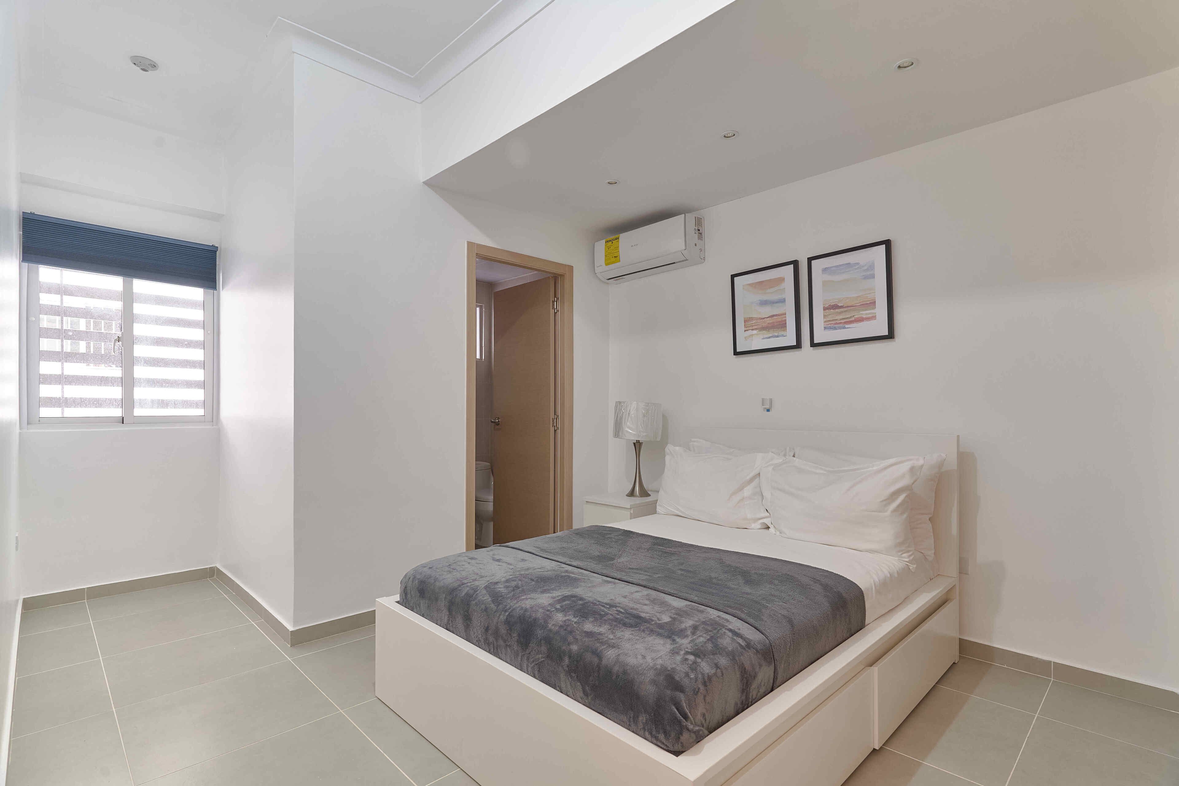Un dormitorio con cama, una ventana con persianas, una puerta, un equipo de aire acondicionado y dos cuadros en la pared.