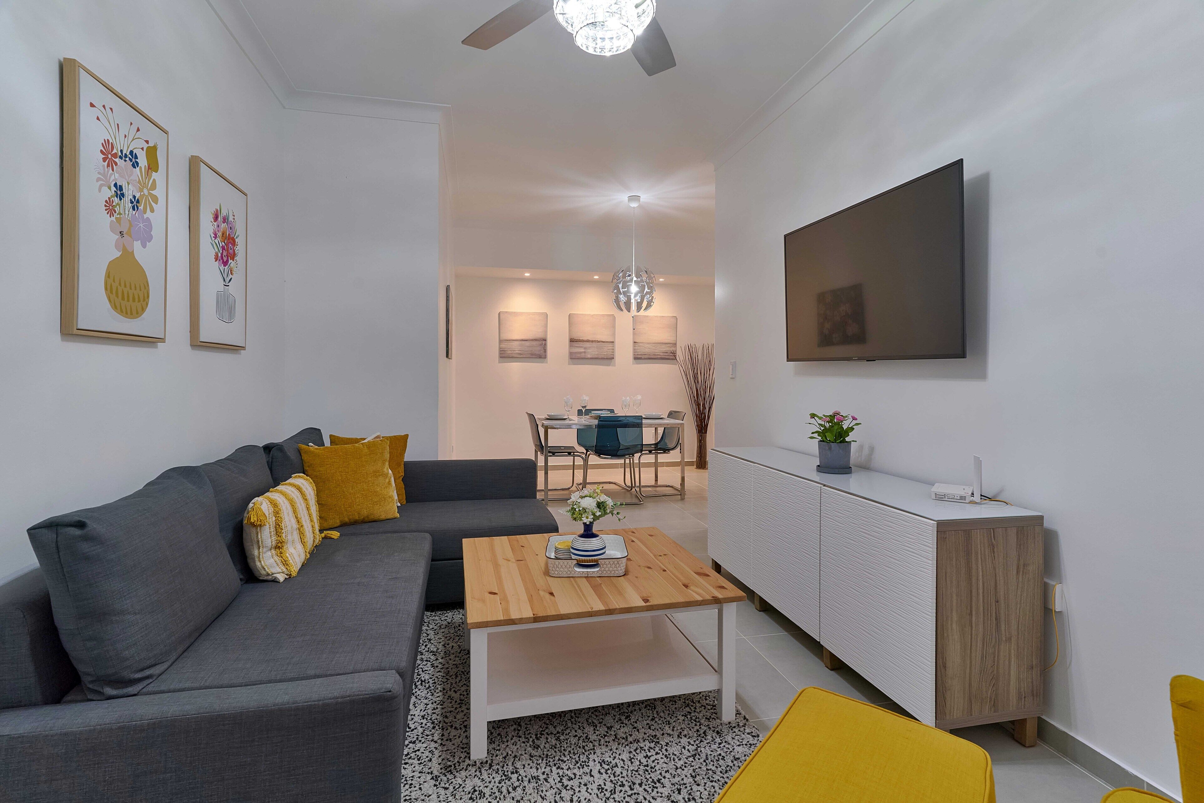 A modern living room with a grey sofa, a wooden coffee table, and a dining area in the background.