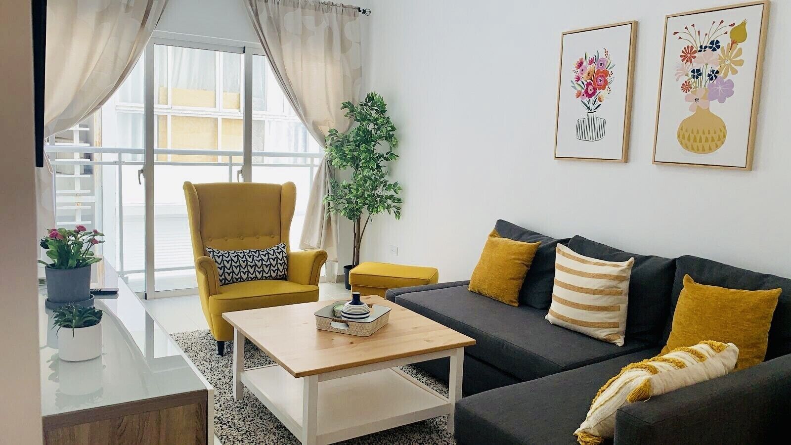 Un salon moderne avec un canapé, un fauteuil et une table basse. Deux œuvres d’art encadrées ornent le mur, et une plante en pot est également présente.