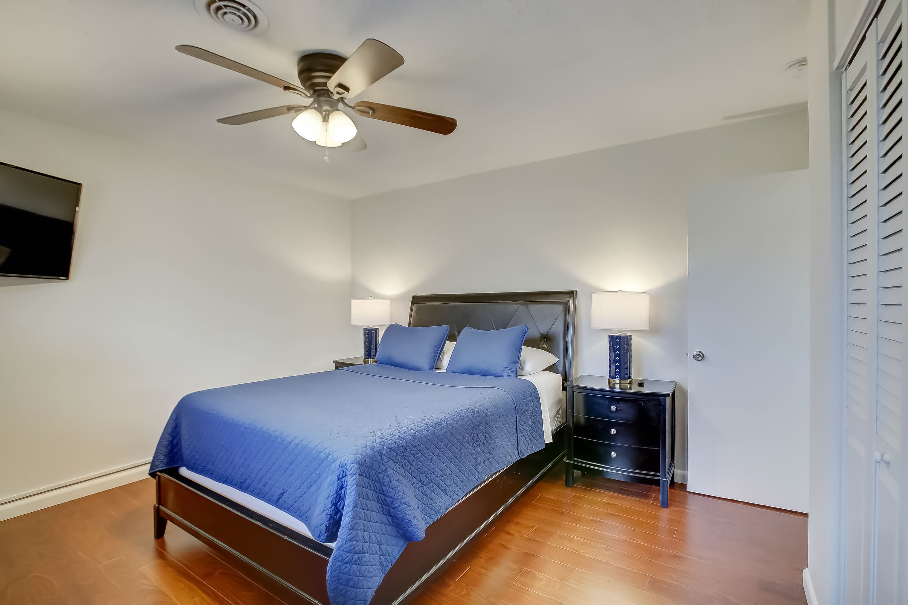 A bedroom with a bed, nightstands, a ceiling fan, and a TV mounted on the wall.