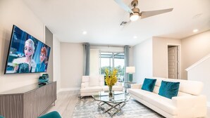 Un salon moderne doté d’un téléviseur à écran plat, de canapés blancs, d’une table basse en verre, de fauteuils turquoise et d’une grande fenêtre agrémentée de rideaux.