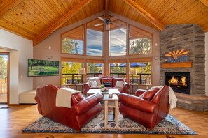 Ein geräumiges Wohnzimmer mit Kamin aus Stein, Ledersofas, einem großen Fernseher und Blick ins Freie.